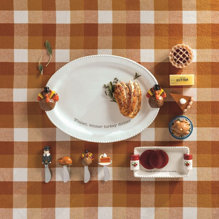 A styled autumn tabletop scene on an orange-and-white checkered tablecloth, featuring a white oval platter with a baked good, small decorative figures, a round tray, a white woven basket-like dish, and other fall-themed serving pieces arranged neatly.