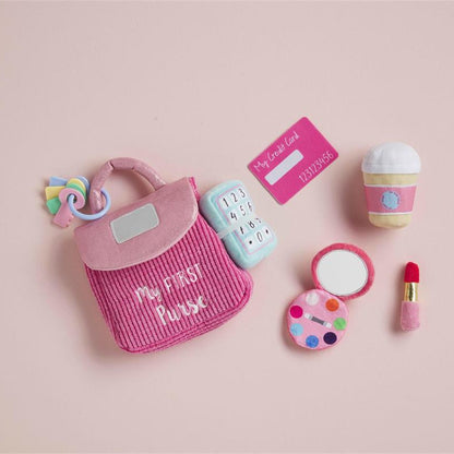 A miniature pink fabric playset featuring a striped handbag with 'My First Playset' embroidered on it, a toy phone, a toy coffee cup, a toy compact mirror, a toy paint palette, and a toy lipstick, arranged on a solid pale pink background.