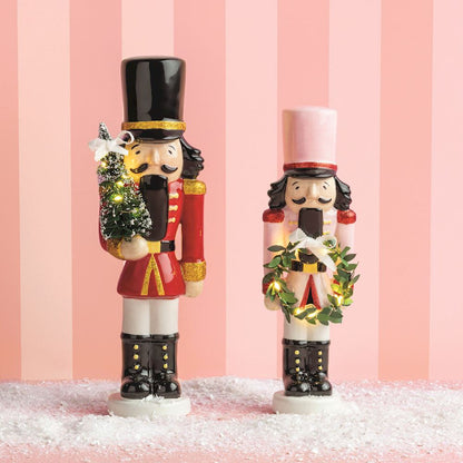 Two decorative nutcracker figurines standing on a white surface with faux snow, set against a pink striped background. One nutcracker wears a red coat and black hat while holding a small green tree, and the other wears a white coat with a pink hat and a green wreath.