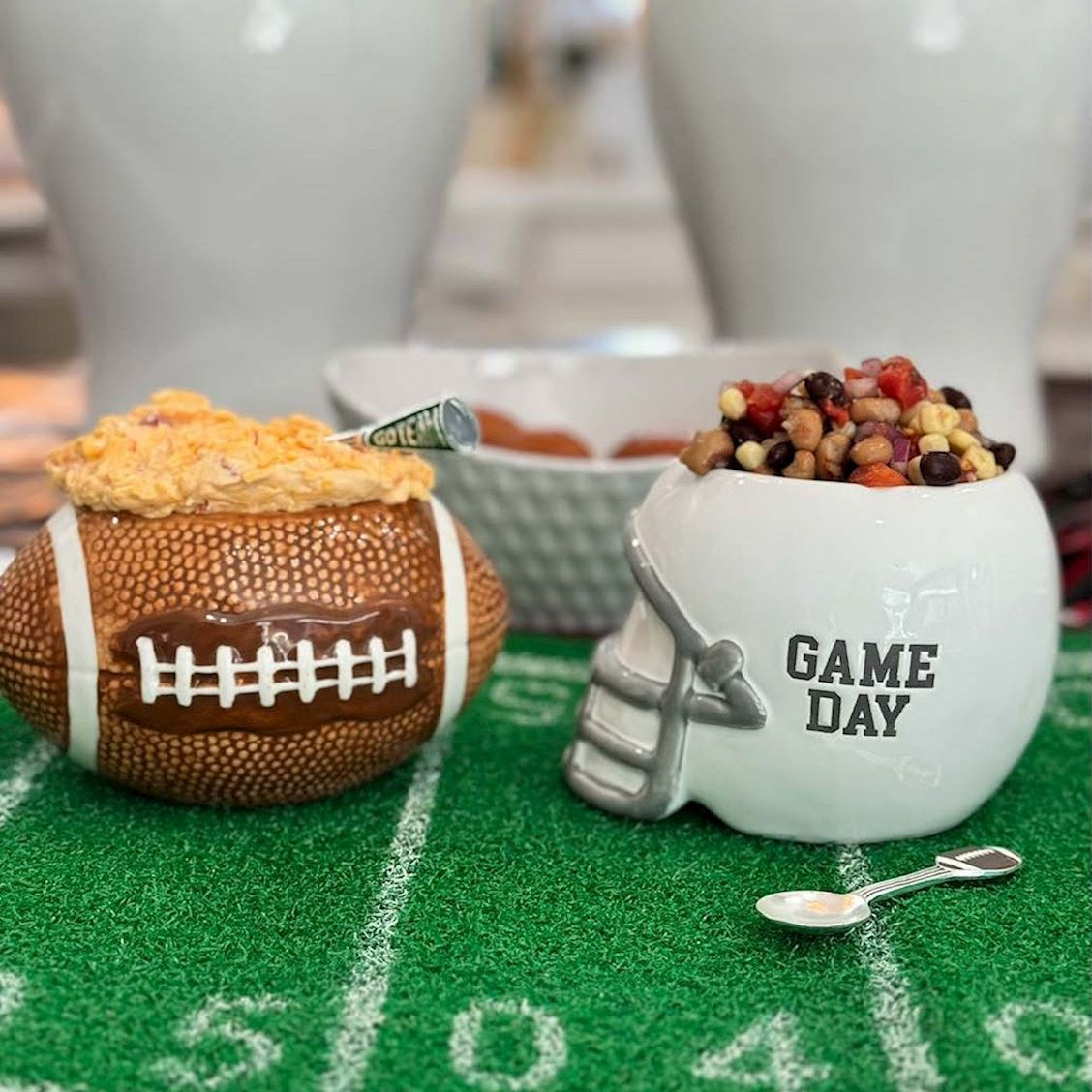 Football-themed party decorations and serving dishes on a green turf mat, including a brown football-shaped bowl with cheese and a white helmet-shaped bowl with beans and the words 'GAME DAY'.
