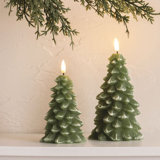 Two green Christmas tree-shaped candles on a white surface with a light beige wall and greenery in the background.