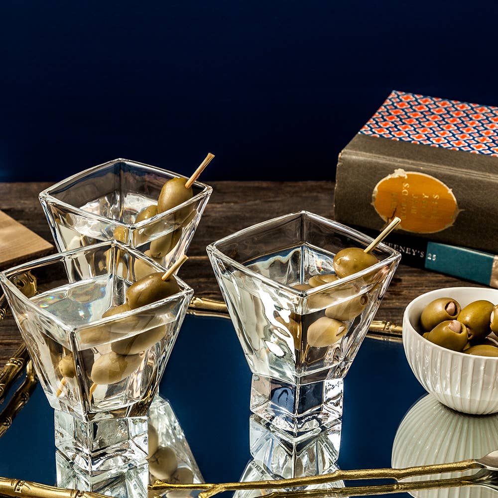 Three square martini glasses with olives on skewers, a bowl of olives, and books in the background.