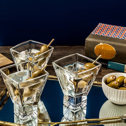 Three square martini glasses with olives on skewers, a bowl of olives, and books in the background.