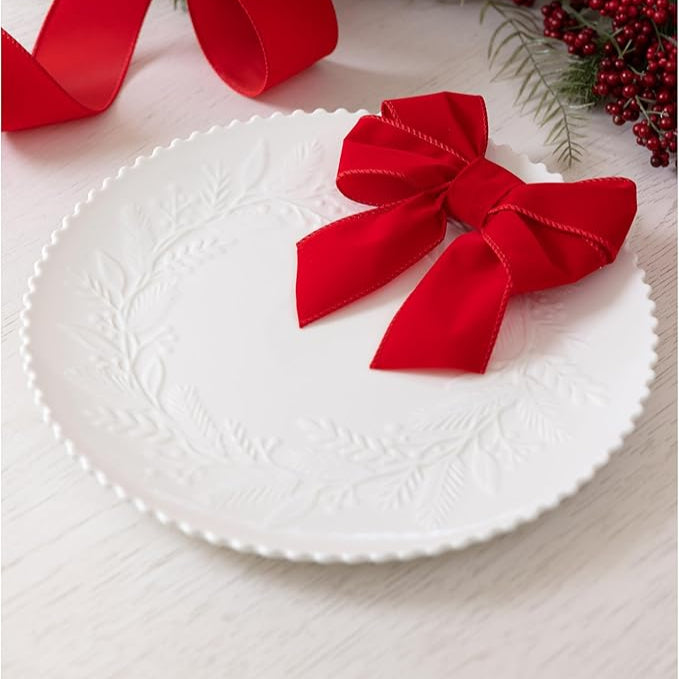 White plate with a red ribbon bow on a white surface with greenery and berries in the background