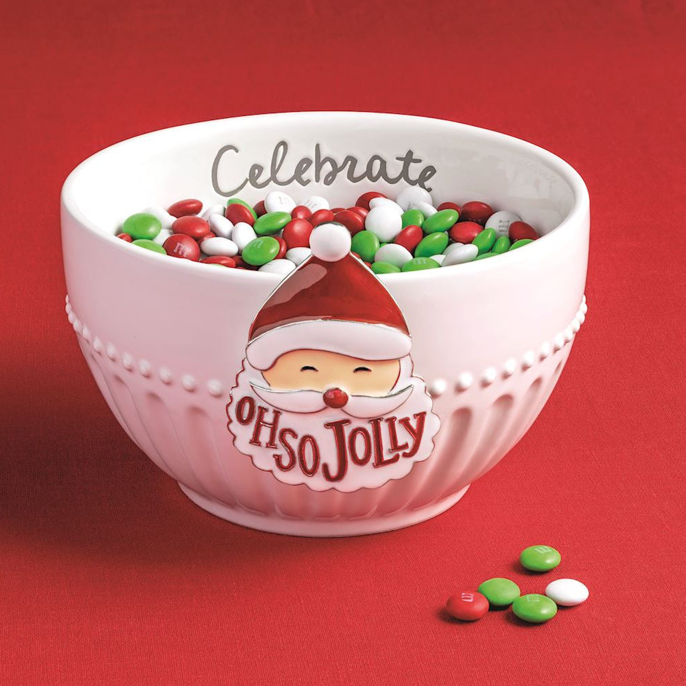 A pink ceramic Christmas bowl filled with red, green, and white candy, featuring 'Celebrate' on the inside rim and a Santa face with 'Oh so Jolly' on the outside. The bowl is set against a solid red background with a few scattered candies in the foreground.