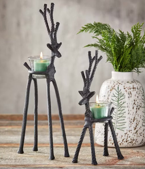 Two black metal reindeer-shaped candle holders on a rustic wooden surface, with green tea light candles in glass holders and a white ceramic vase with greenery in the background.