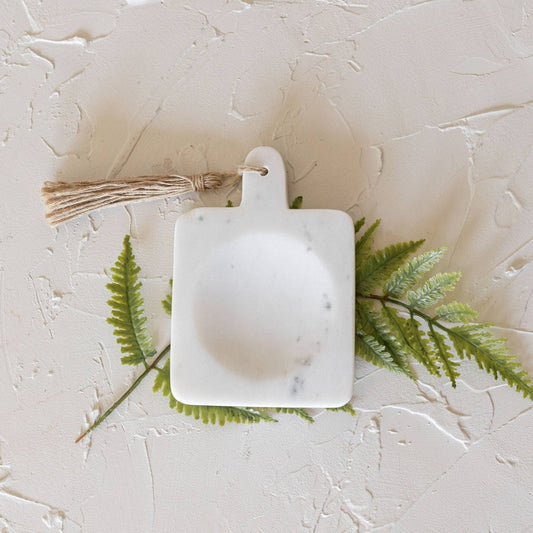 Small white marble cutting board with a tassel on a textured beige surface with green leaves.