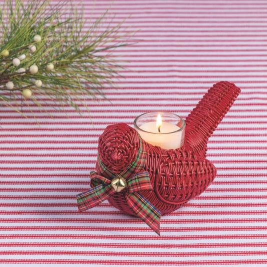 A red woven bird-shaped candle holder with a lit white candle inside, decorated with a red plaid ribbon bow and small gold bell, set on a pink and white striped fabric background with pine branches in the corner.