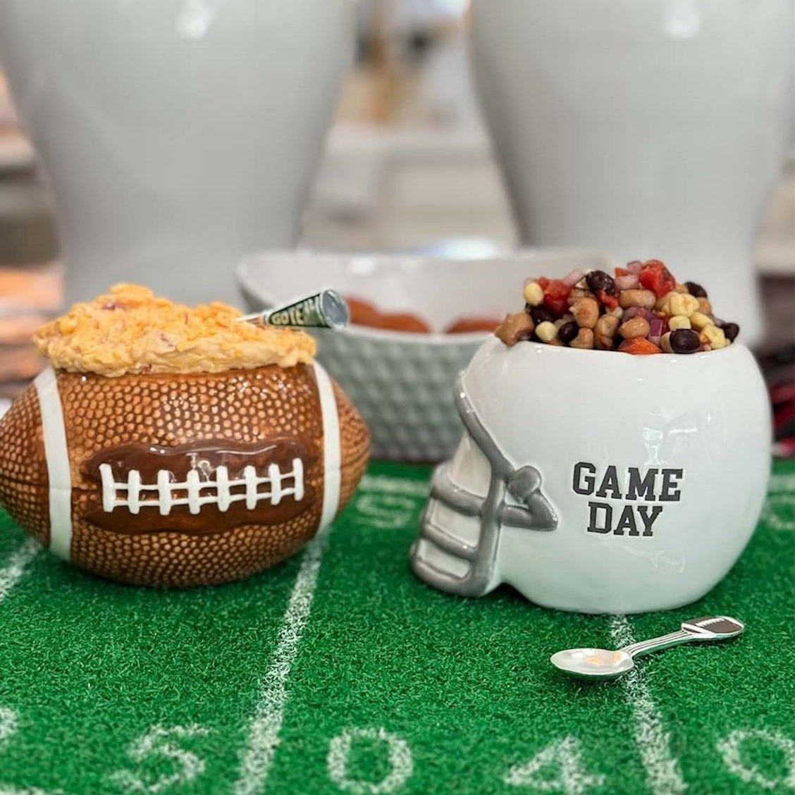 Football-themed party decorations and serving dishes on a green turf mat, including a brown football-shaped bowl with cheese and a white helmet-shaped bowl with beans and the words 'GAME DAY'.