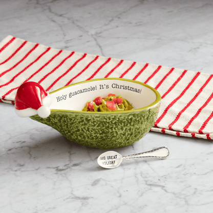 A green textured ceramic bowl with a Santa hat handle and printed text inside, filled with guacamole and a small spoon, styled on a gray marble surface with a red and white striped napkin.
