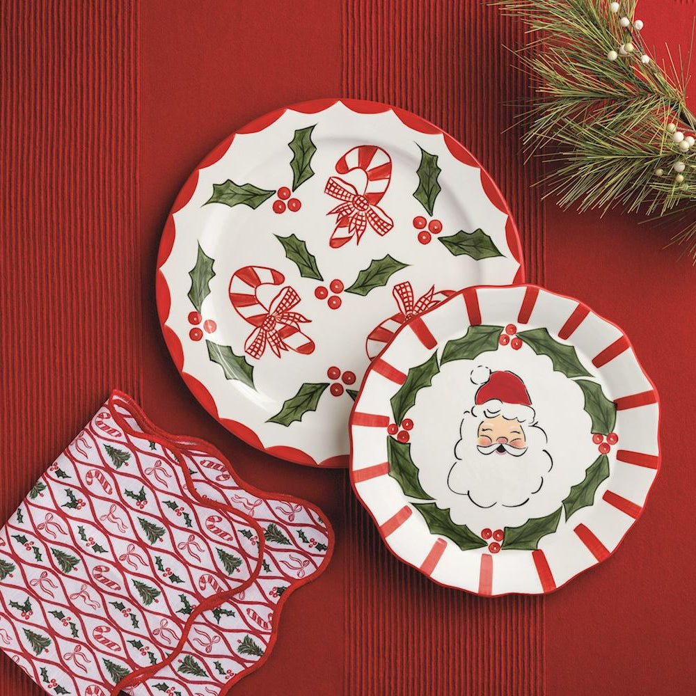 Christmas-themed tableware set featuring two white scalloped plates with red and green holiday patterns, a matching red and white patterned napkin, and a pine branch on a red background.