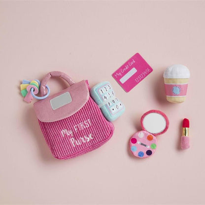 A miniature pink fabric playset featuring a striped handbag with 'My First Playset' embroidered on it, a toy phone, a toy coffee cup, a toy compact mirror, a toy paint palette, and a toy lipstick, arranged on a solid pale pink background.