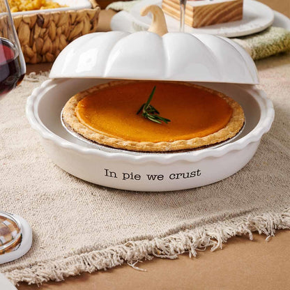 A white ceramic pumpkin-shaped pie dish with a matching lid, shown on a table runner with a pumpkin pie inside. The front of the dish reads 'In pie we crust'.