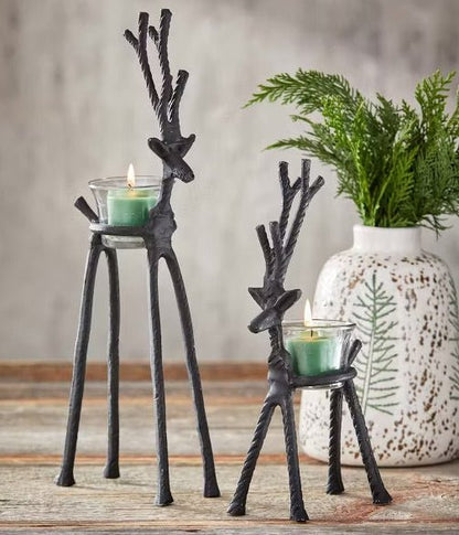 Two black metal reindeer-shaped candle holders on a rustic wooden surface, with green tea light candles in glass holders and a white ceramic vase with greenery in the background.